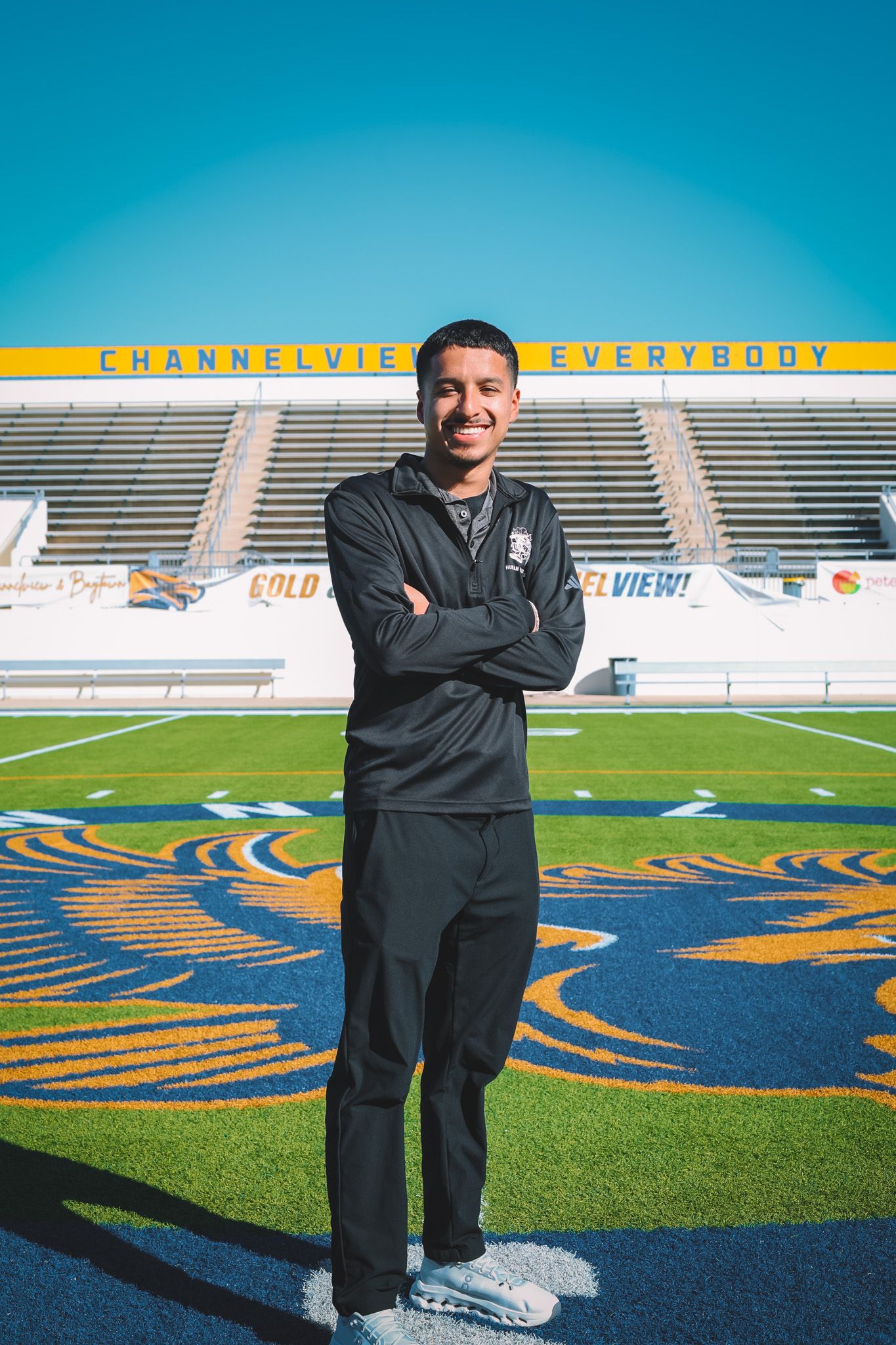 A person in dark clothing stands on a football field with blue and gold markings at a stadium.