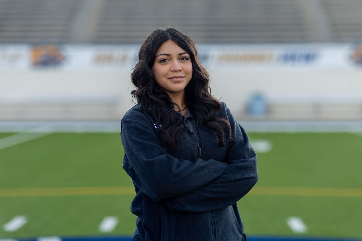 A person in a dark jacket poses with crossed arms on a football field.