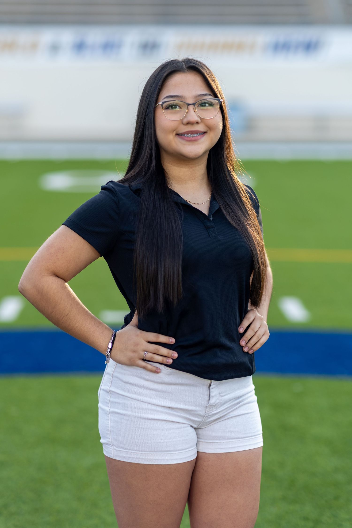 A person in glasses wearing a black polo shirt and white shorts poses on a sports field with artificial turf.
