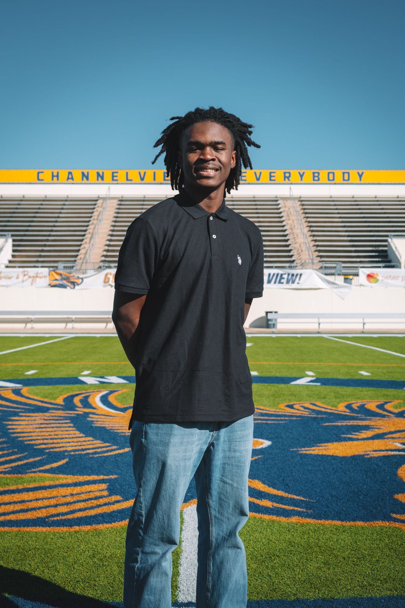A person stands on a football field wearing a black shirt and jeans with stadium seating visible in the background.