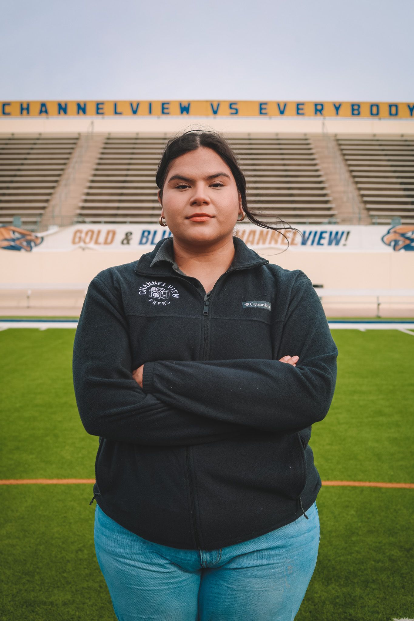 A person in a black Columbia fleece jacket and blue jeans stands with crossed arms on a football field with stadium seating.