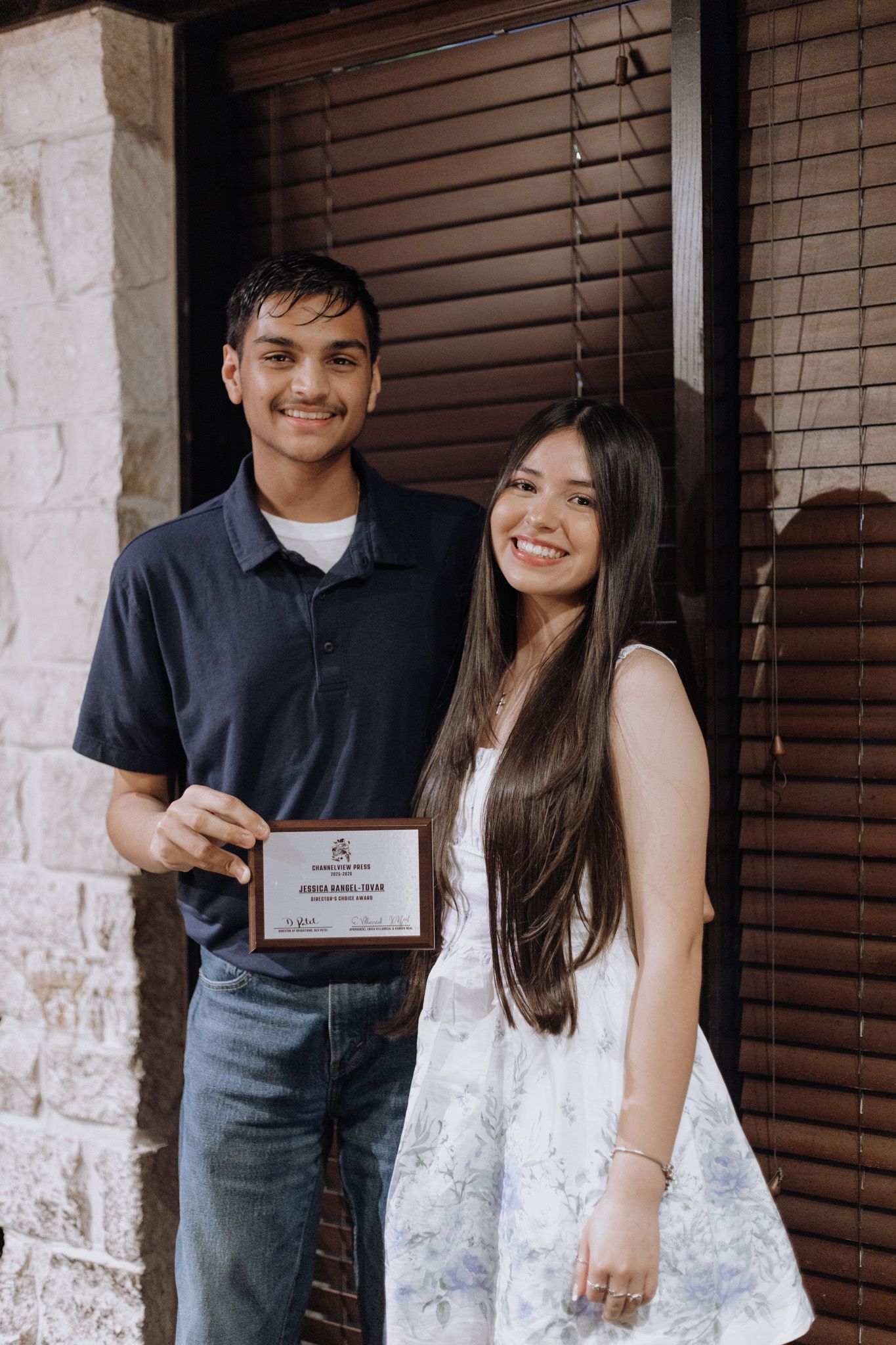 Two young people smiling and holding an award certificate while standing in front of a wooden door.