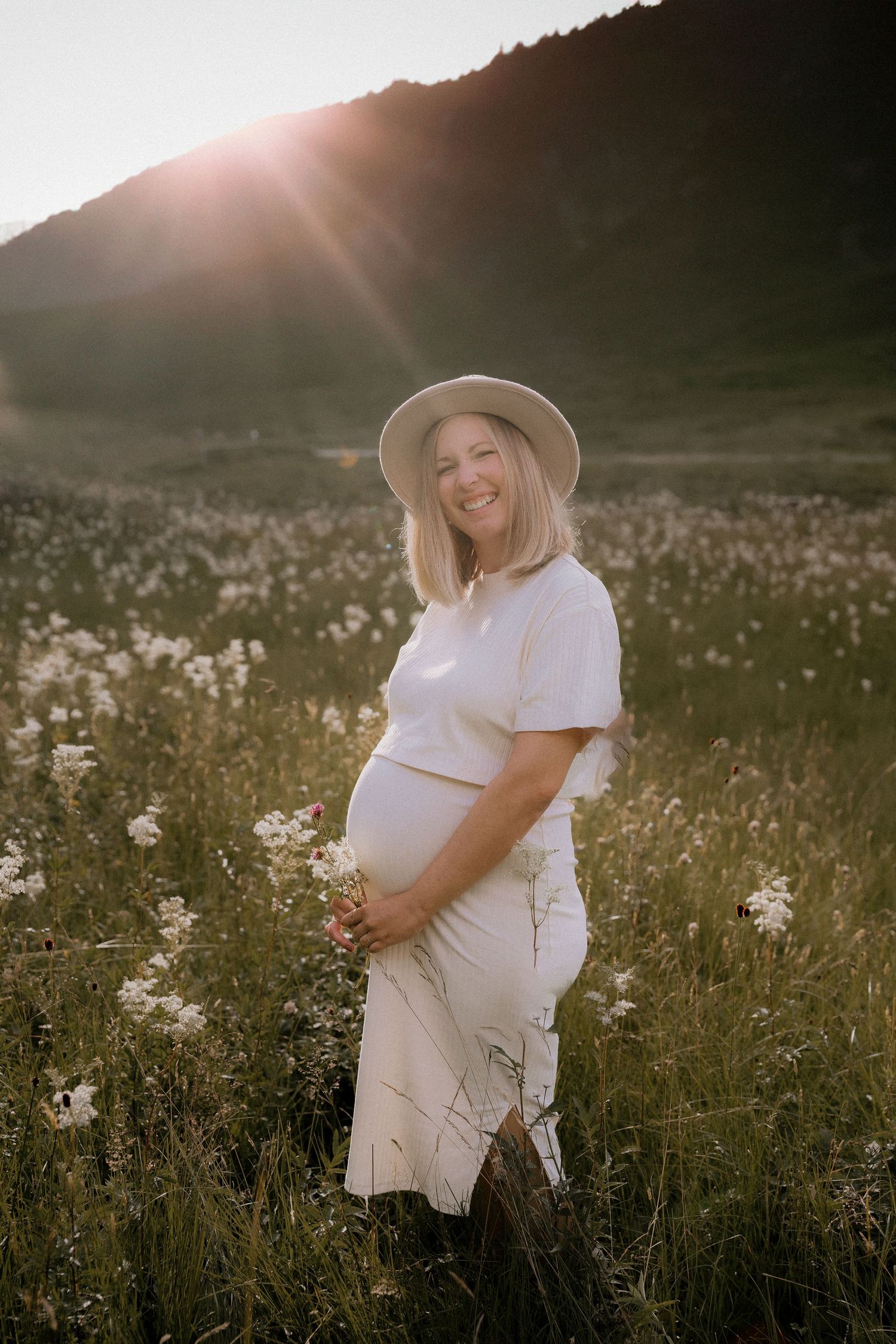 An expectant mother in a white dress and hat poses in a sunny wildflower meadow at sunset.