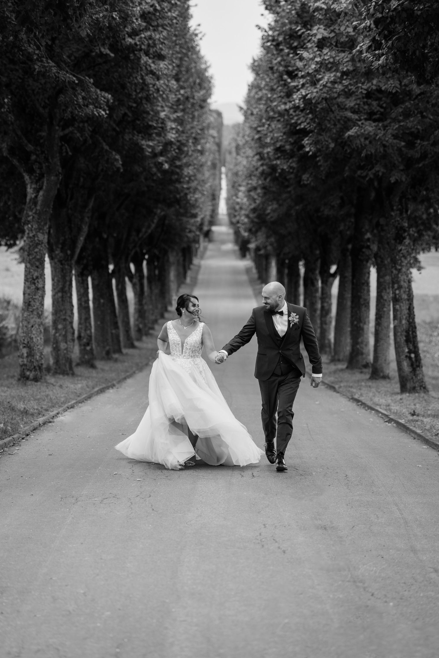 Black and white wedding photo of couple walking down a tree-lined avenue.