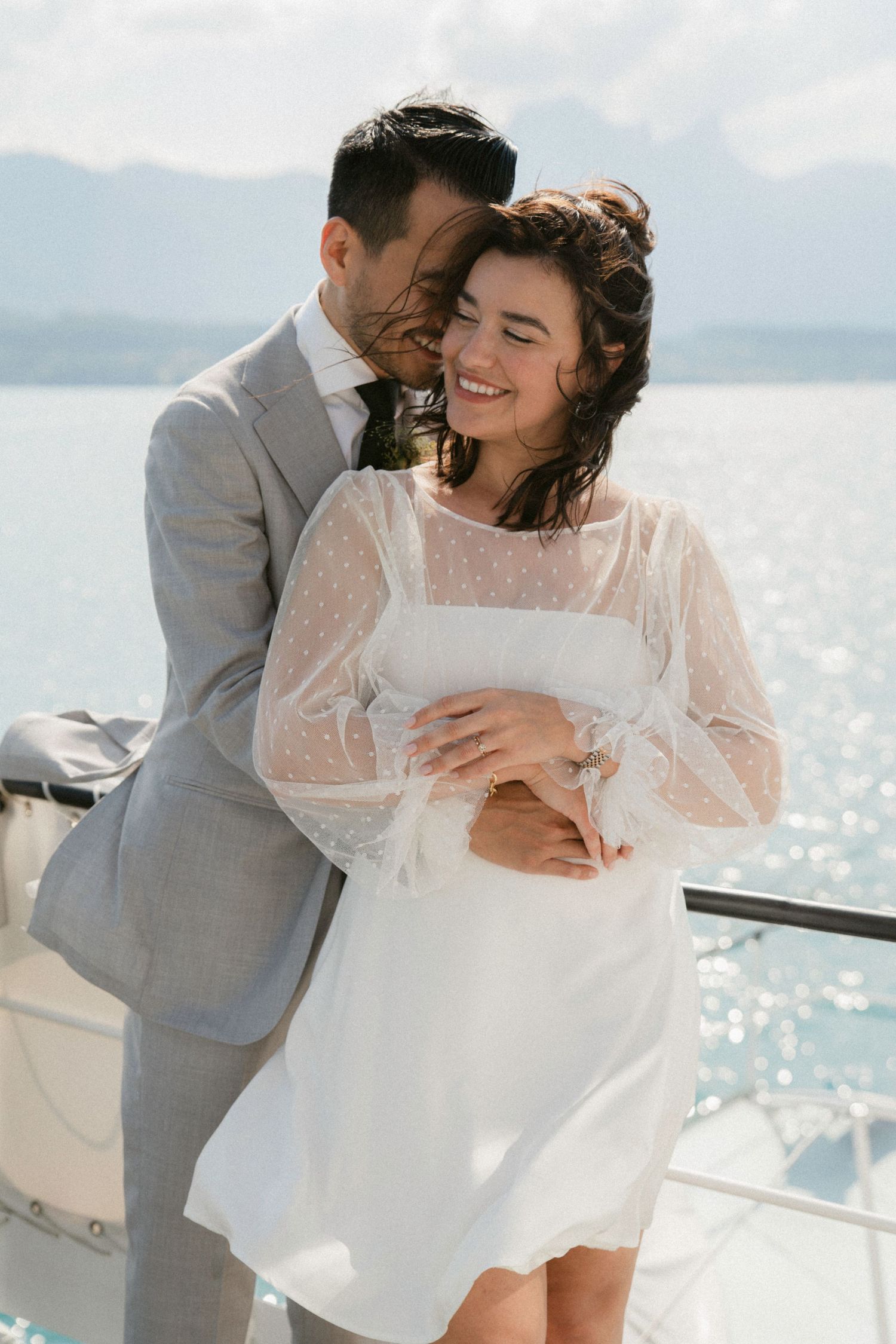 A romantic wedding couple embrace on a boat deck overlooking sparkling water and mountains on a sunny day.