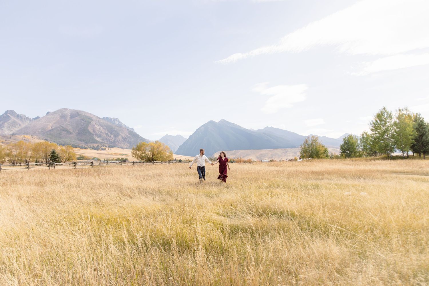 Two figures walking through golden grass fields with majestic mountains and autumn trees in the background on a sunny day.