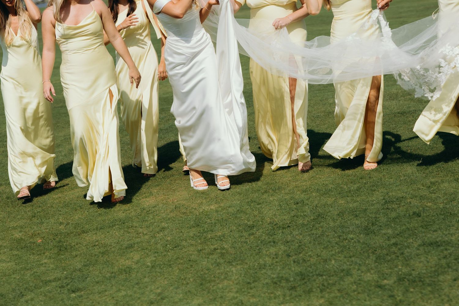 Wedding party in flowing white and cream dresses walking together on green grass lawn.