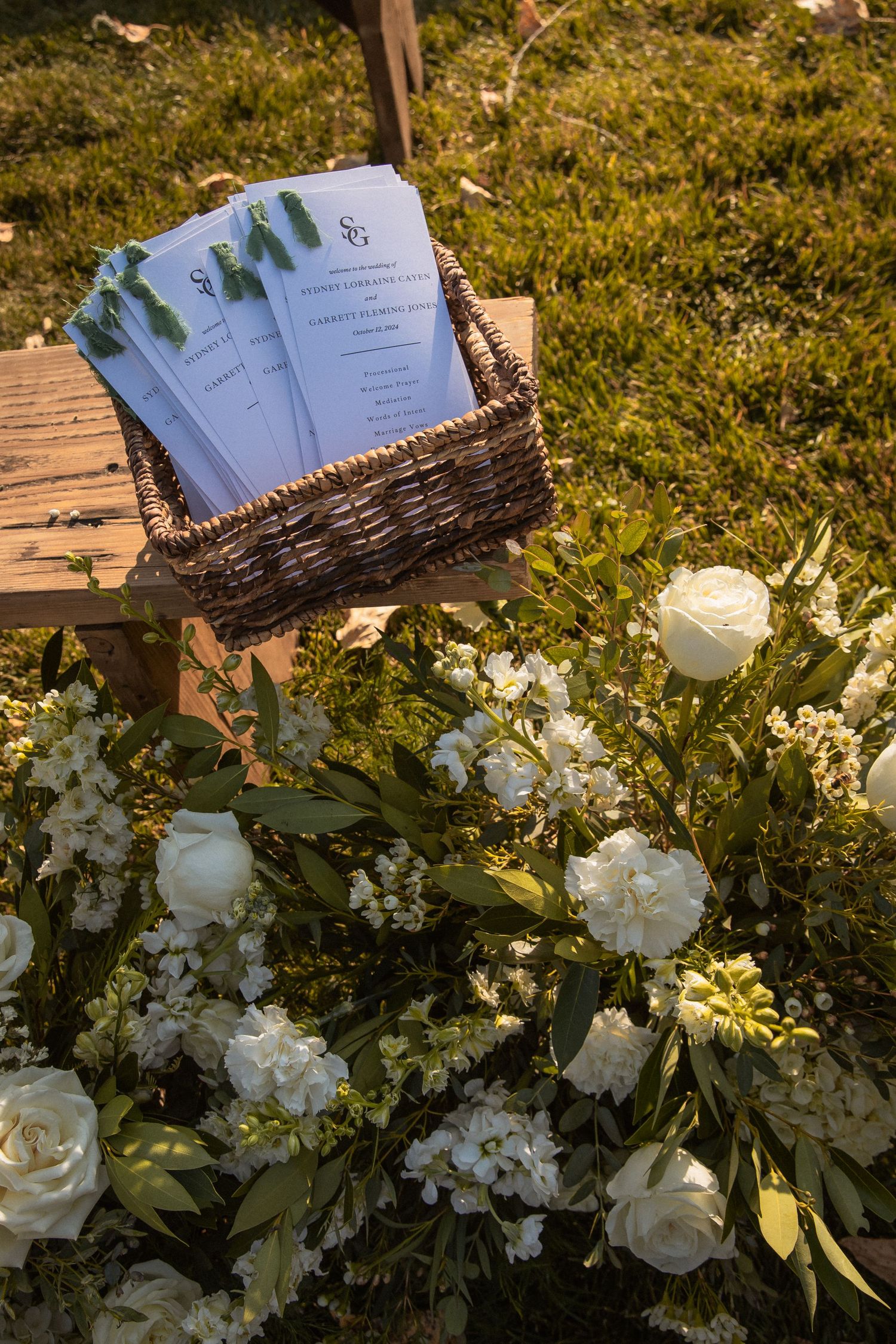 Wedding ceremony programs in wicker basket next to white roses and floral arrangements on grassy lawn.