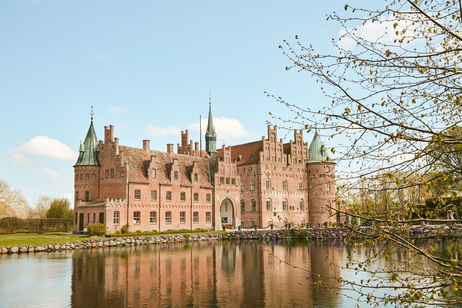 A grand European castle with pink stone walls reflects in the surrounding moat on a sunny day.