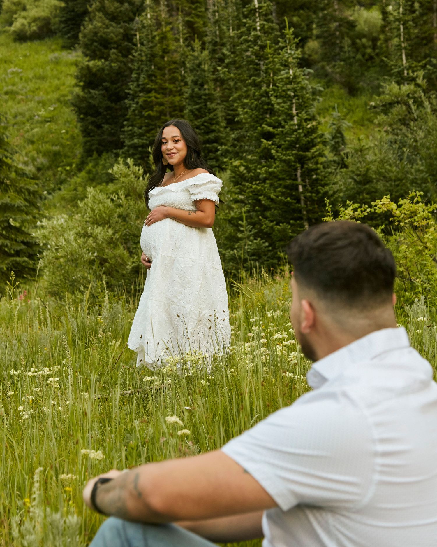 Maternity photo in mountain meadow with person in white dress while partner looks on from foreground.