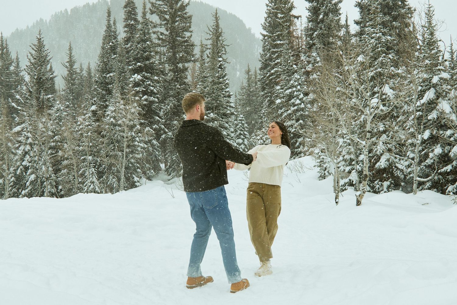 Two people walk together through a snowy winter landscape surrounded by tall evergreen trees in the mountains.