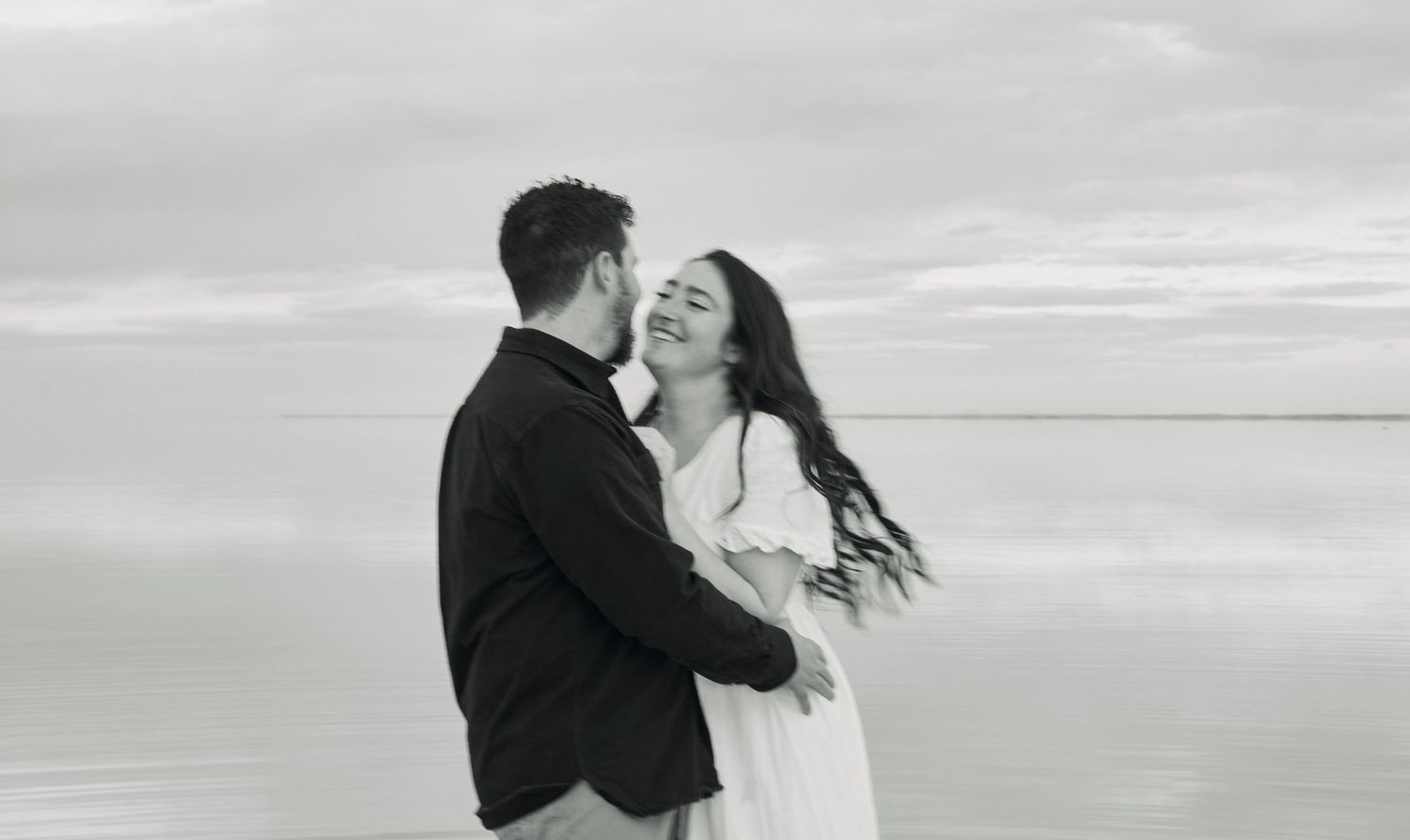 Black and white photo of a joyful couple laughing together against a waterfront backdrop.