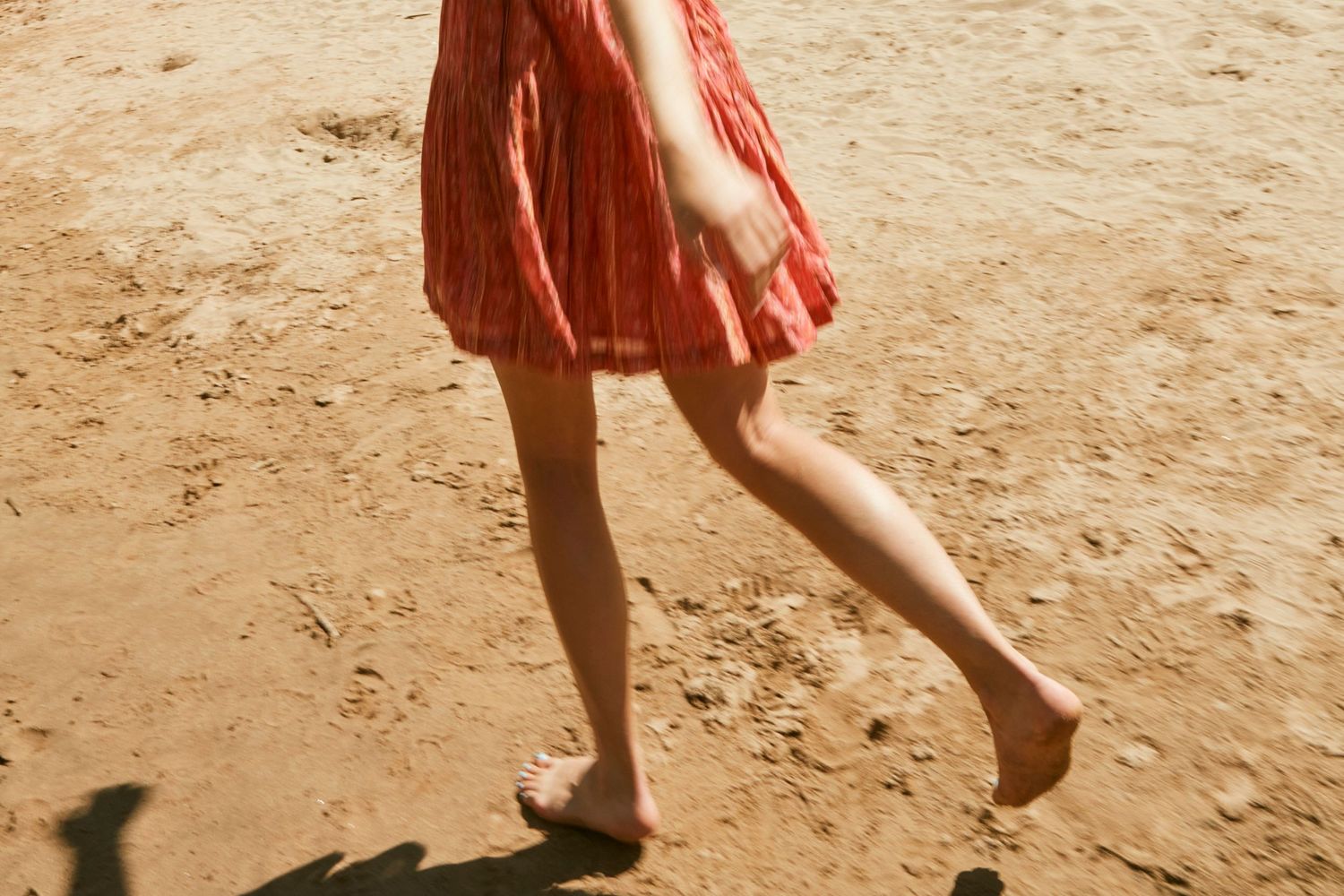 Barefoot person in flowing coral dress walking on sandy terrain.