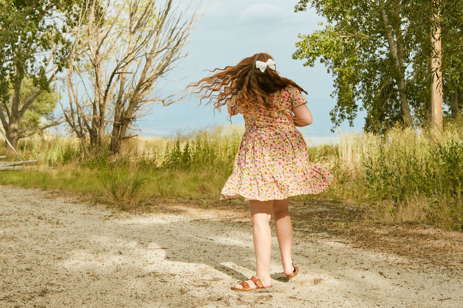 A girl in a floral dress twirls on a dirt path surrounded by trees and grass on a her birthday.