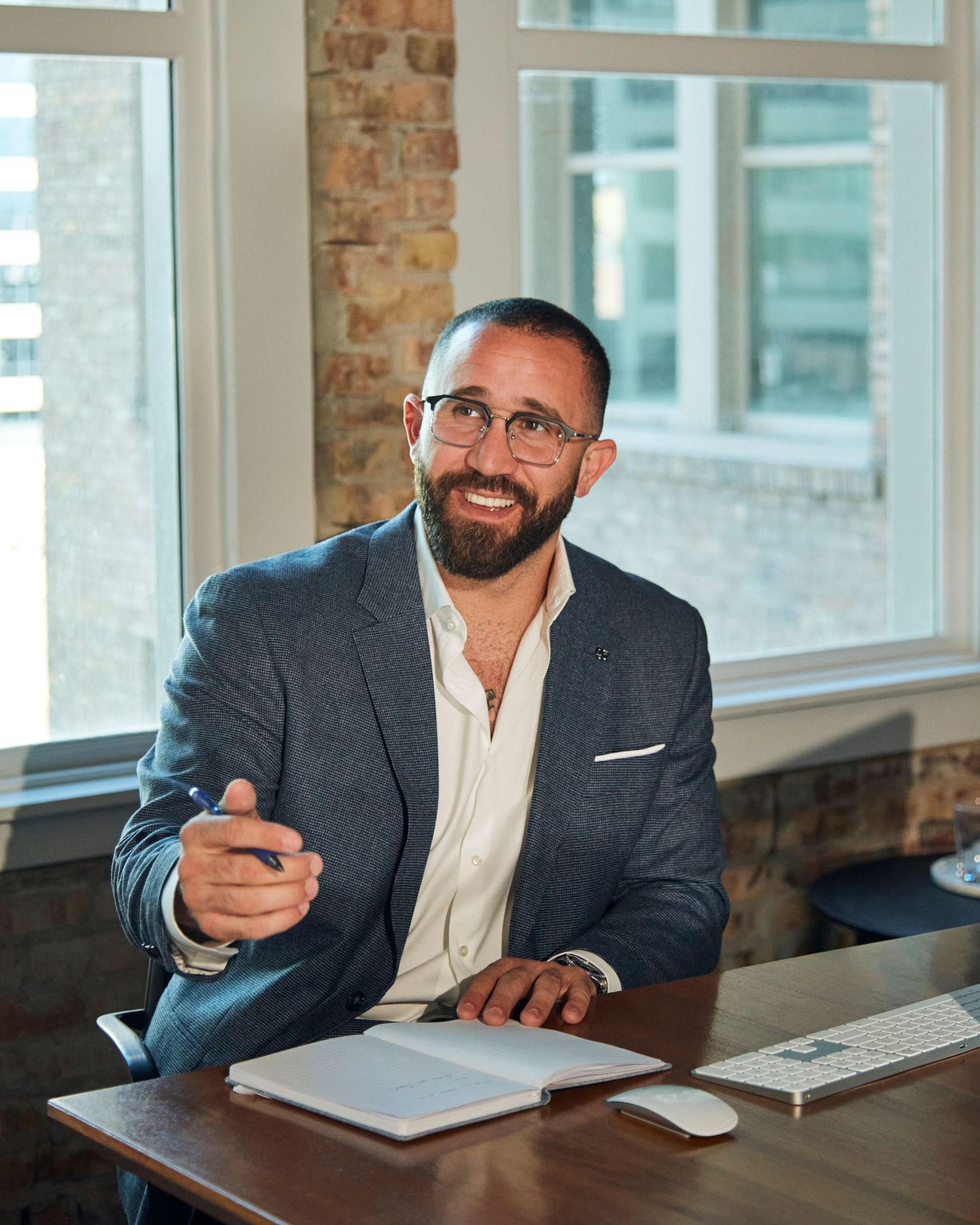 Professional in gray blazer works at modern desk with Apple computer and city views.