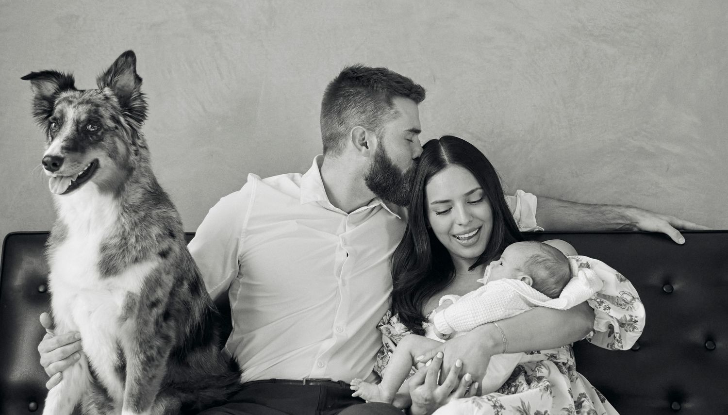 Family relaxes on leather couch with newborn baby and Australian shepherd dog in black and white photo.