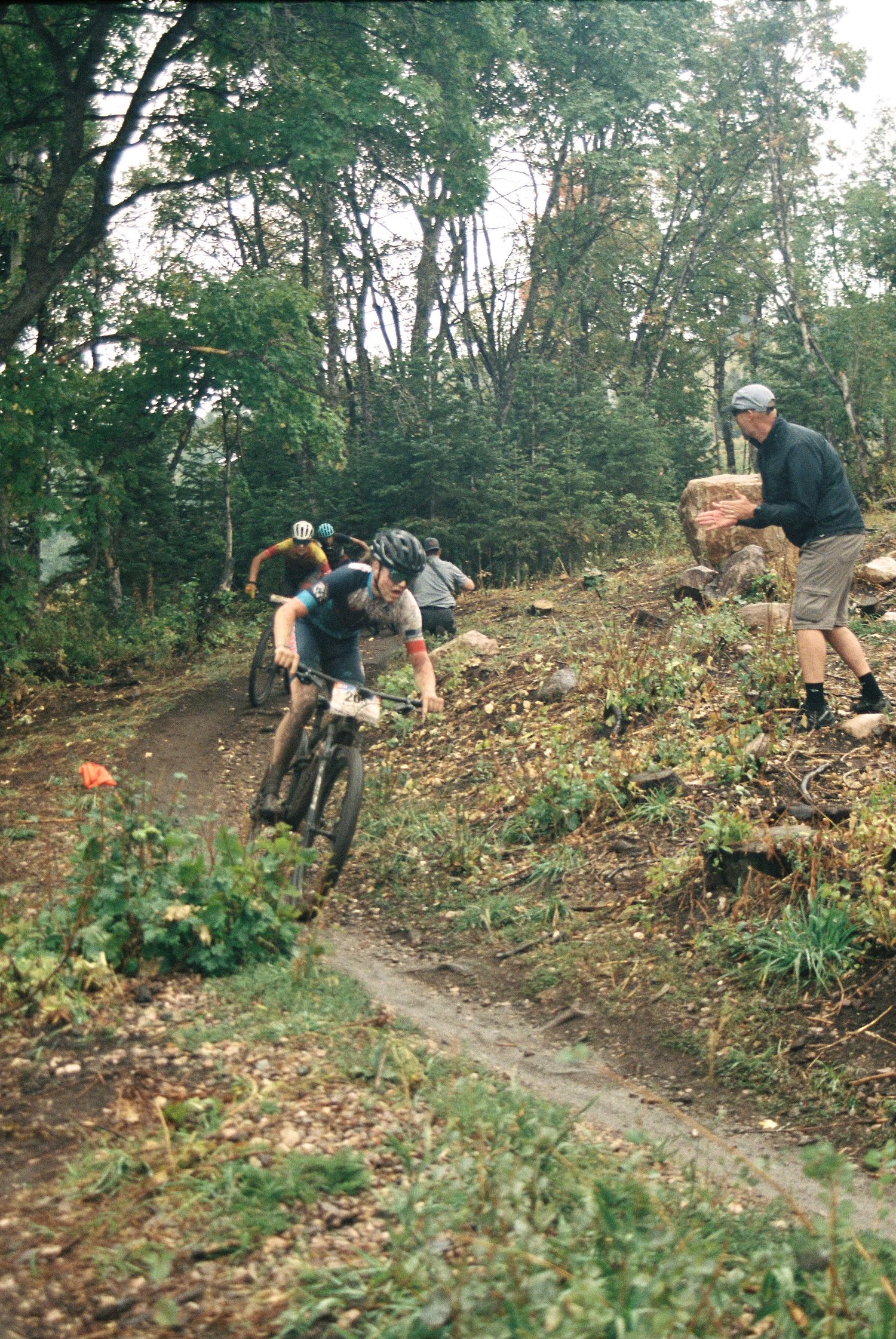 Mountain bikers navigating a steep dirt trail while spectator watches from sideline.