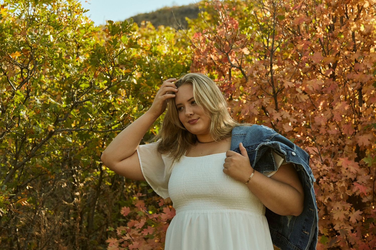 Person in white dress with denim jacket poses against fall colors.