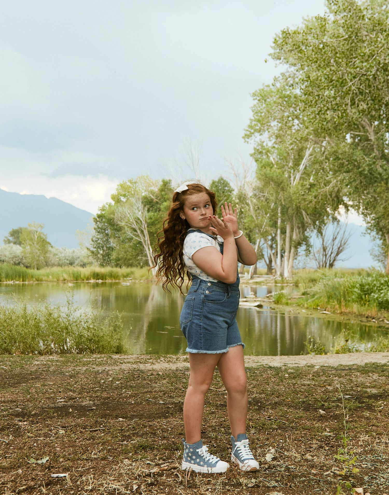 Young person in denim overalls poses playfully by a scenic mountain lake.