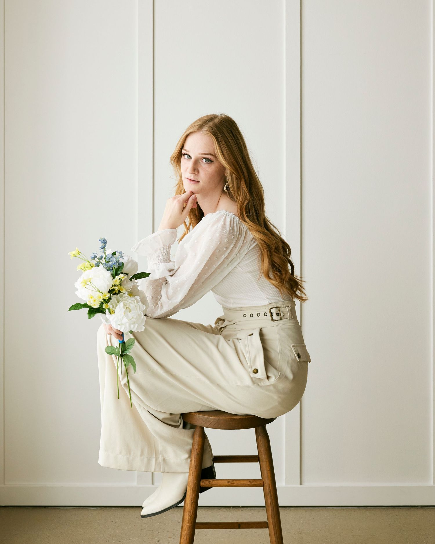 Person in white outfit poses gracefully on wooden stool with flowers against minimalist white wall.