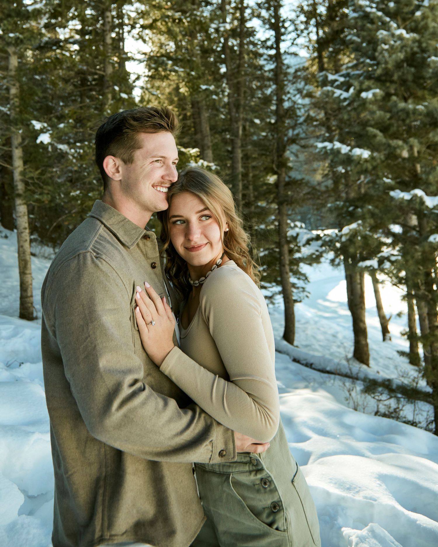 Intimate winter engagement photo of couple embracing in snowy forest setting.