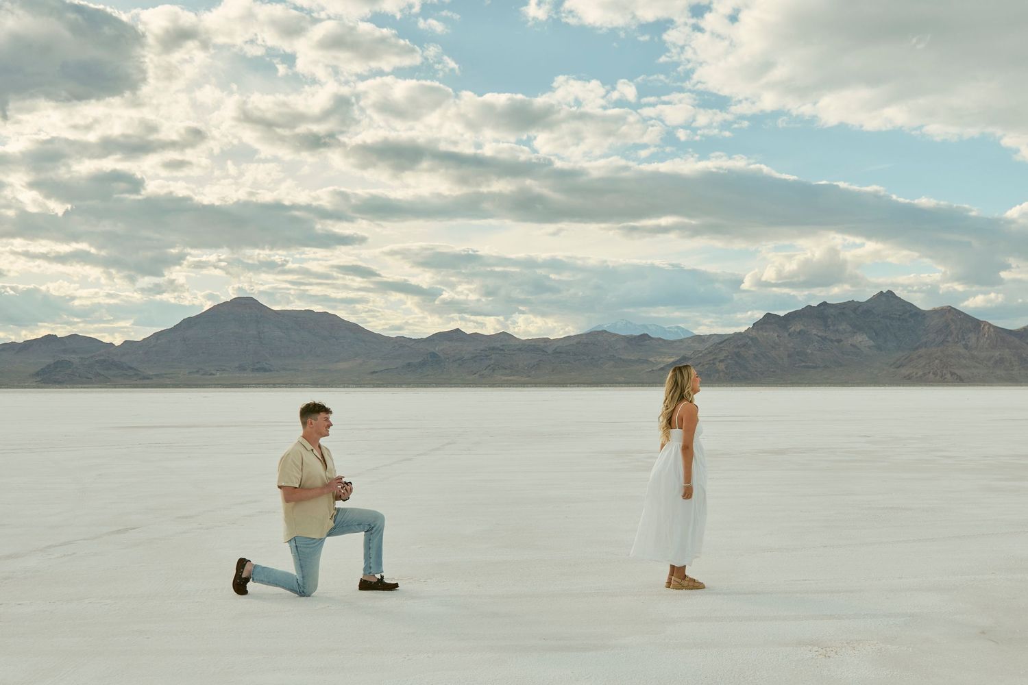 A marriage proposal taking place on a white salt flat with mountains in the background at sunset.