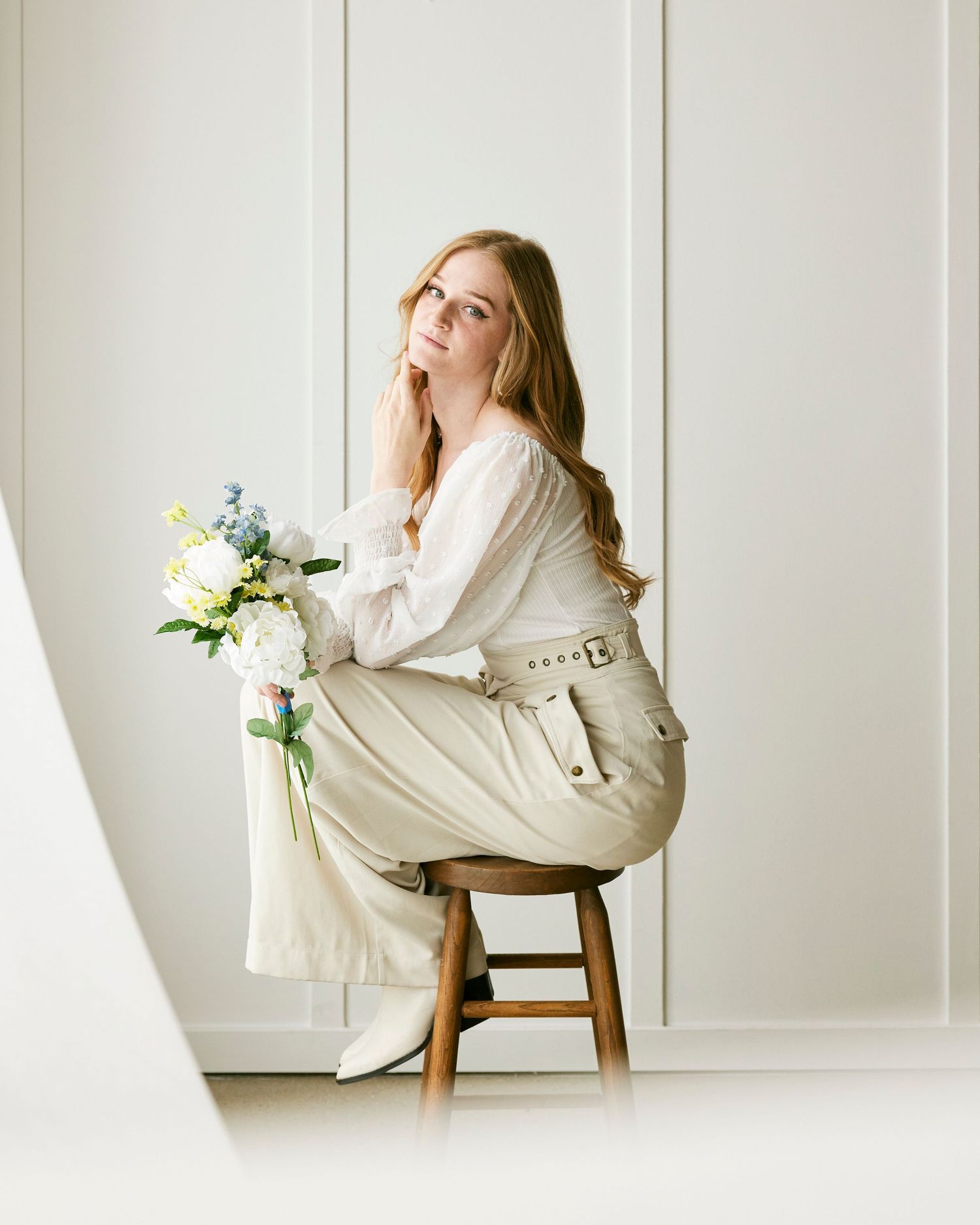 Soft indoor portrait with white flowers and crisp natural lighting against white wall.