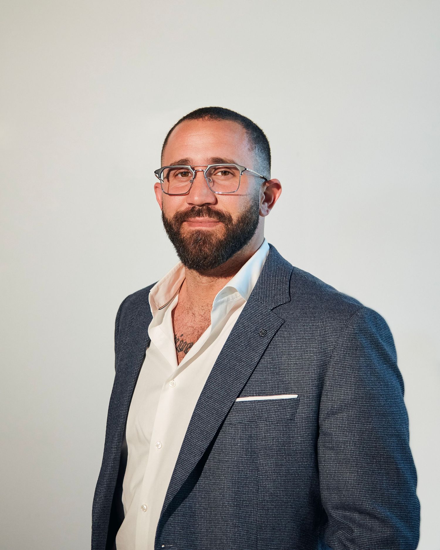 Professional in gray suit and glasses poses against plain wall in modern office setting.