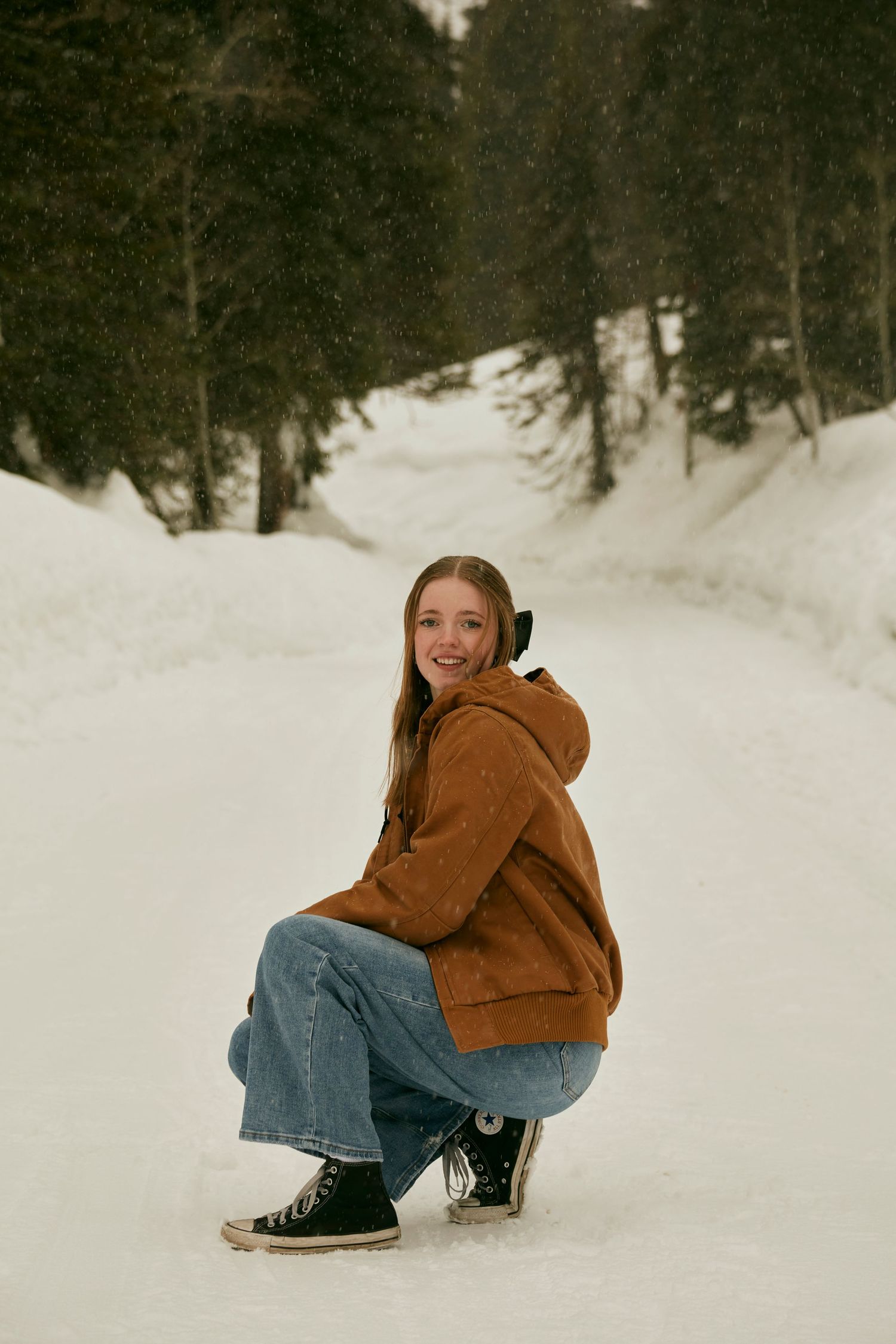 A winter scene showing someone crouching in the snow wearing a brown hoodie and jeans.
