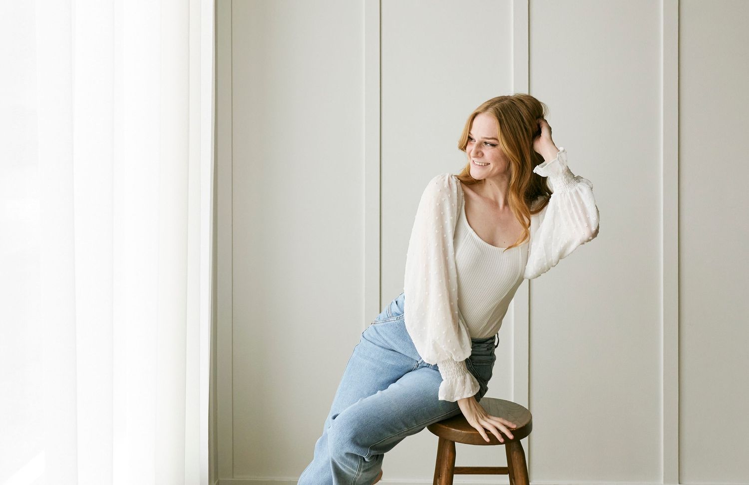 Casual lifestyle portrait on wooden stool wearing jeans and light sweater against white wall.