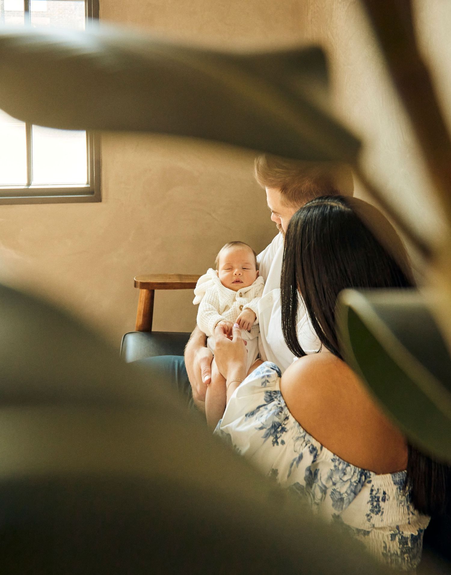 Through foliage, a glimpse of a baby in white being cradled on a wooden chair.