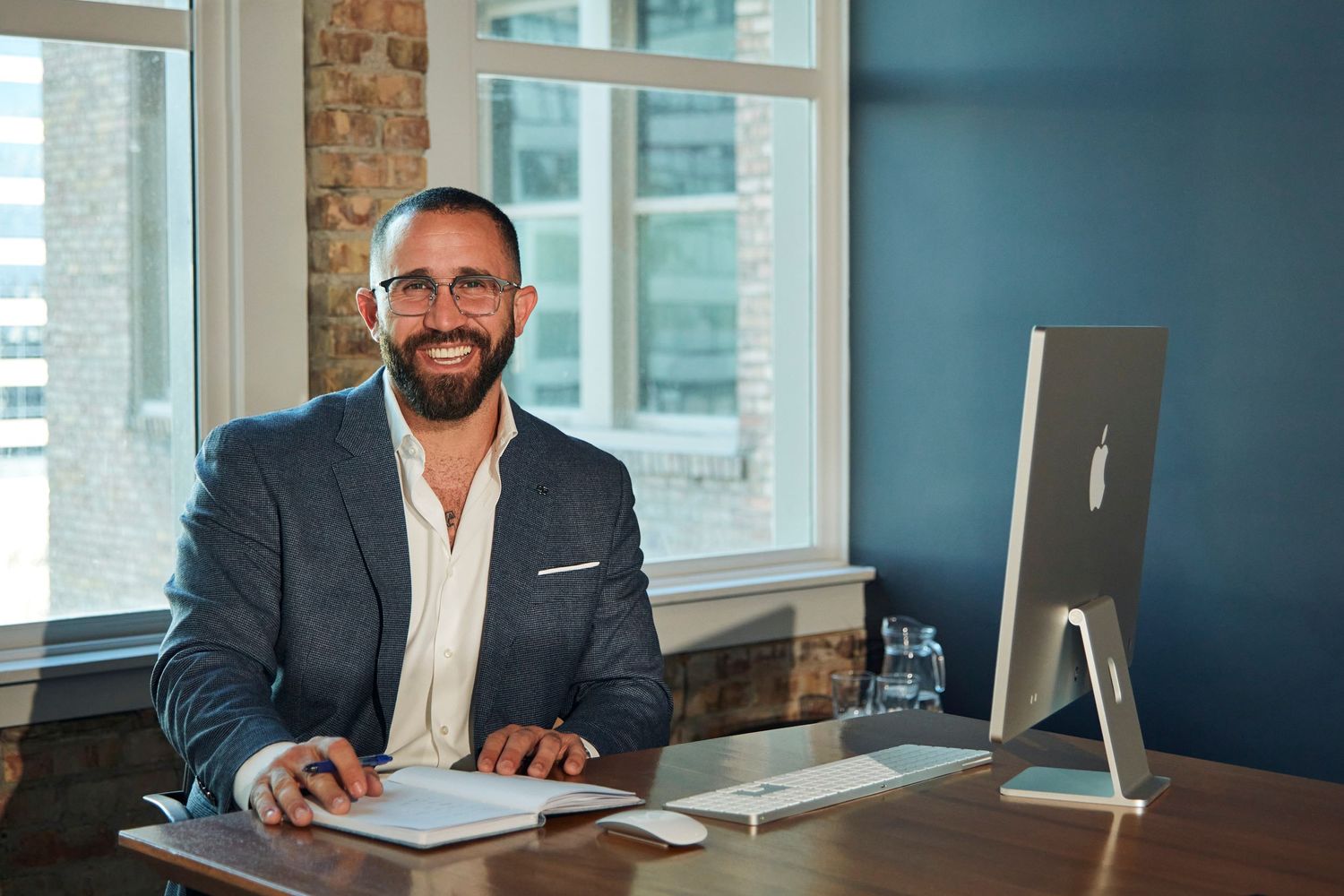 Business professional works at modern desk with iMac display and exposed brick office walls.