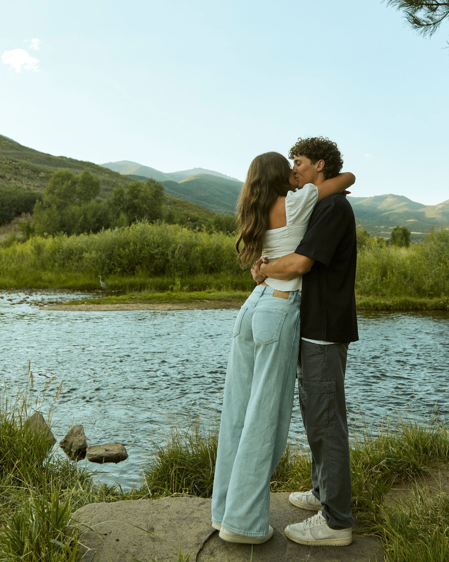 Romantic couple standing by river with mountain vista in background.