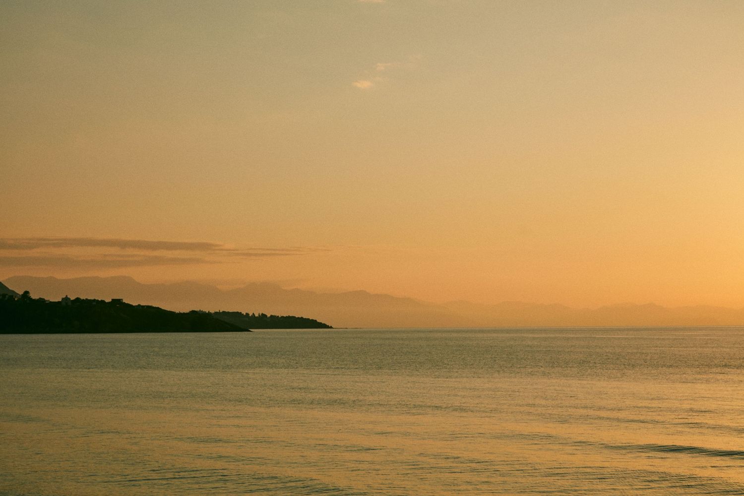 Serene ocean sunset with golden sky reflecting on calm waters near a coastal silhouette.