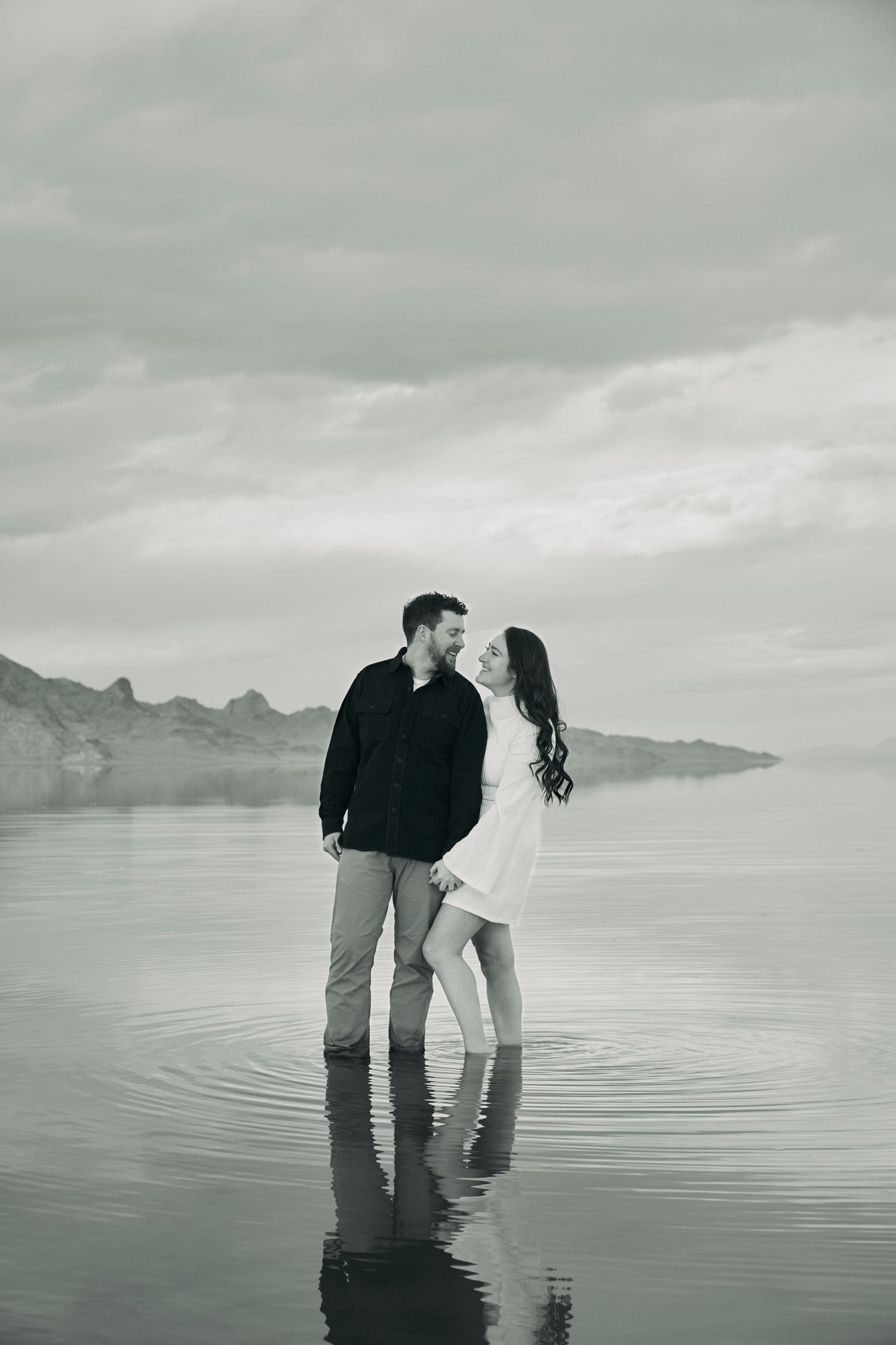 Black and white photo of a couple standing in reflective water with mountains in background.