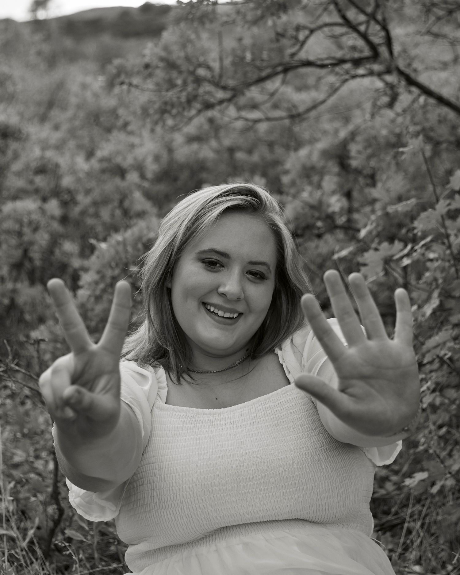 Person wearing white sweater making peace sign and showing palm against blurred nature background.