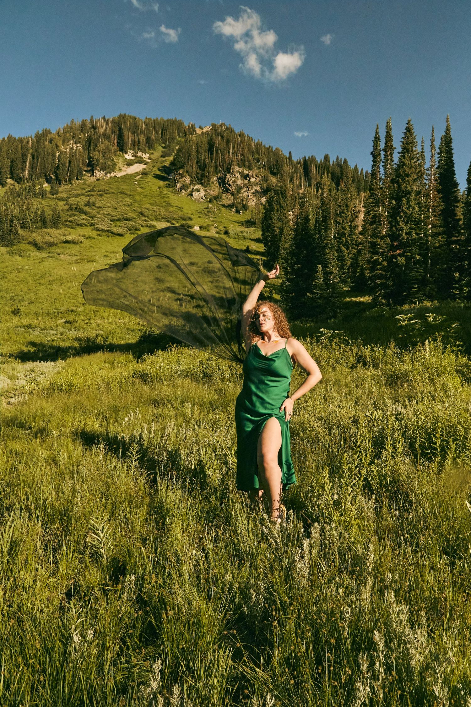 Dancing figure in green dress on mountainside meadow with evergreen forest backdrop.