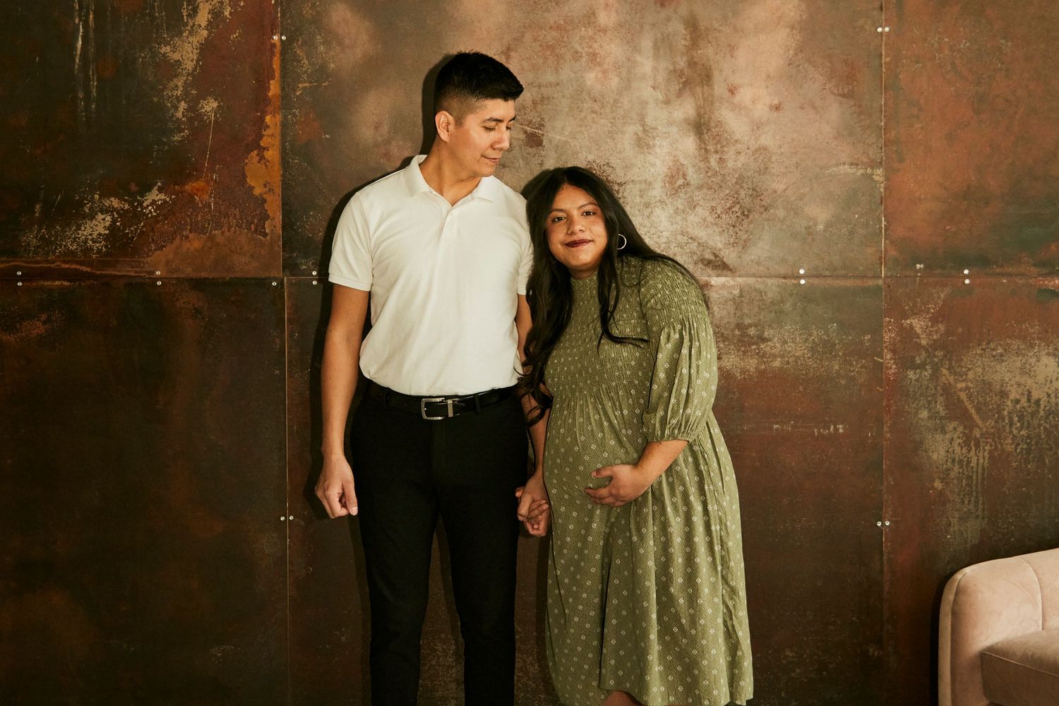 A couple poses together against a rustic metal wall during a maternity photoshoot.