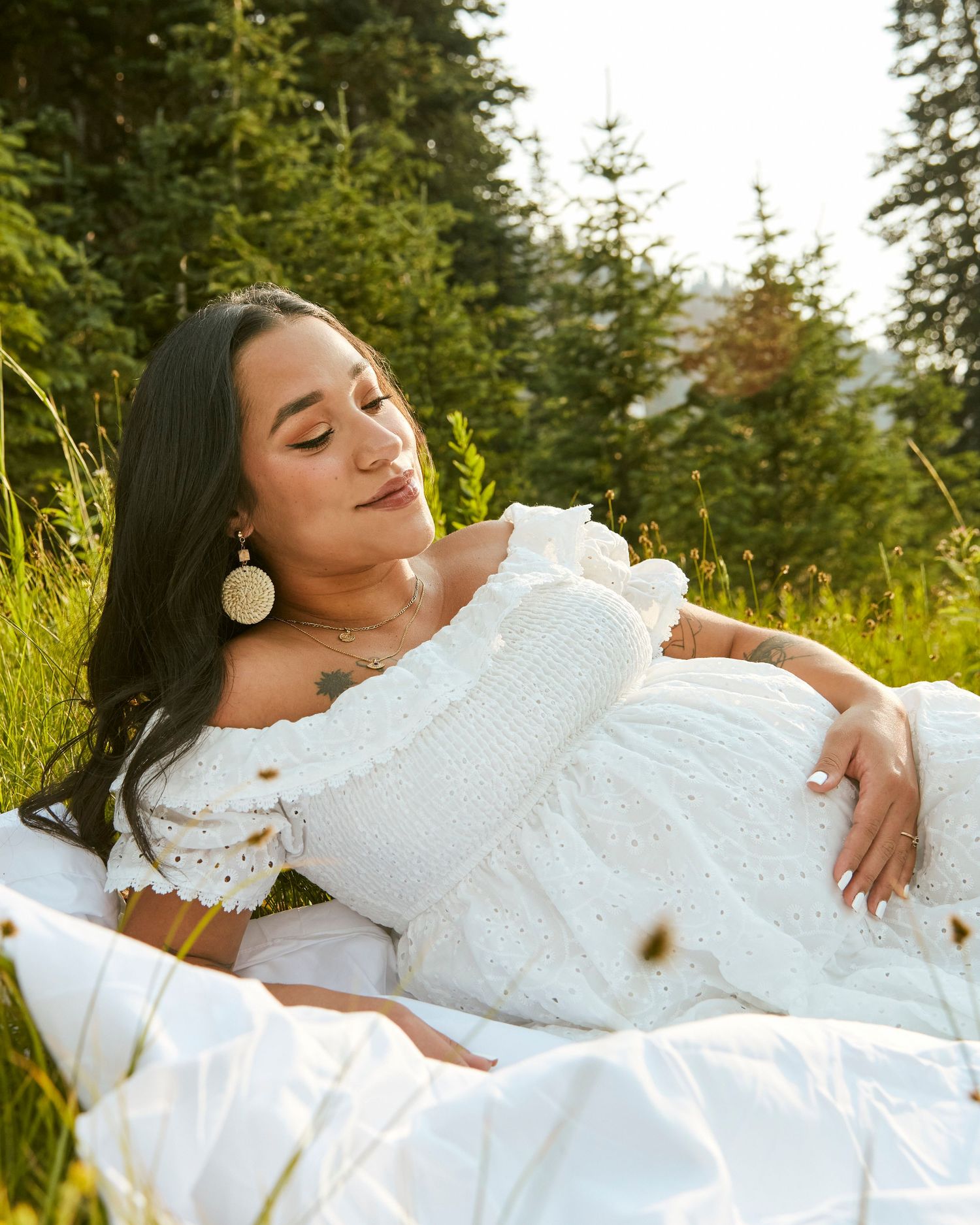 Serene maternity portrait in a white dress laying on a blanket in a sunlit mountain meadow.