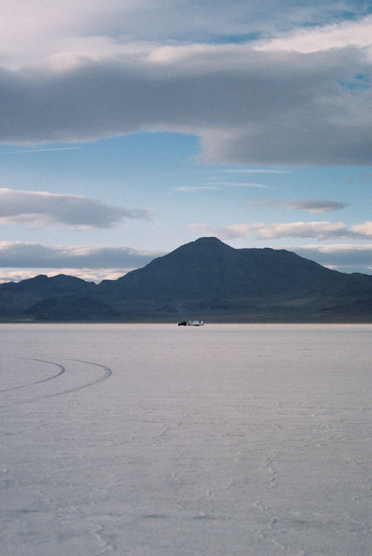 Tire tracks curve across Bonneville Salt Flats with mountains silhouetted against cloudy sky.