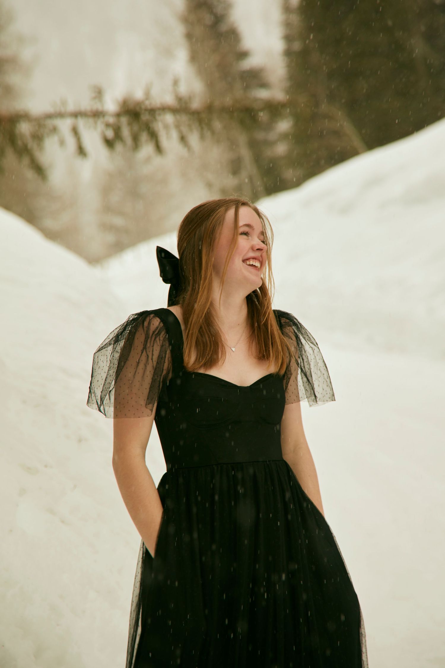 A woman in a black tulle dress poses joyfully in a snowy winter landscape.
