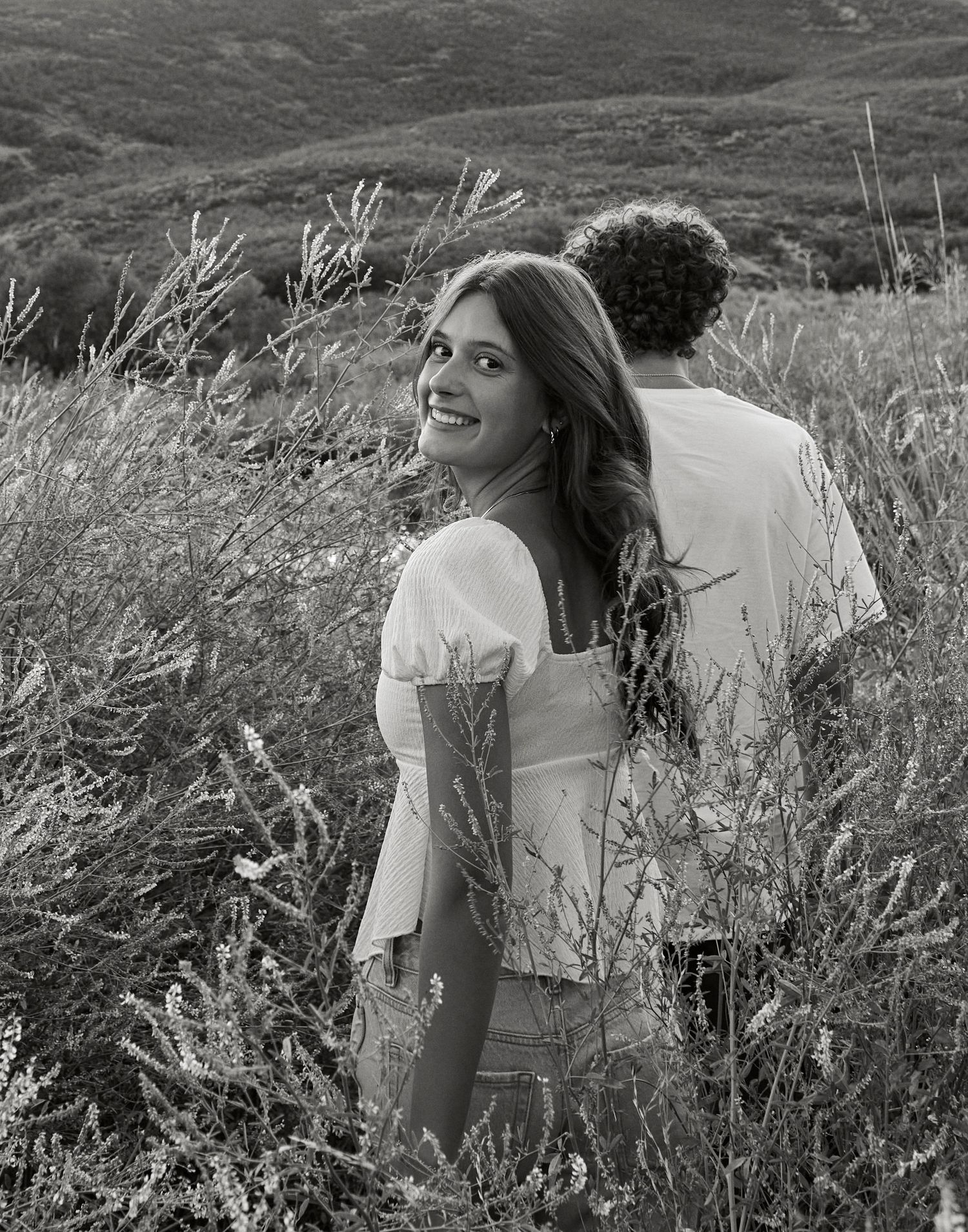 Couple sharing a romantic moment in tall grass against a mountainous landscape.