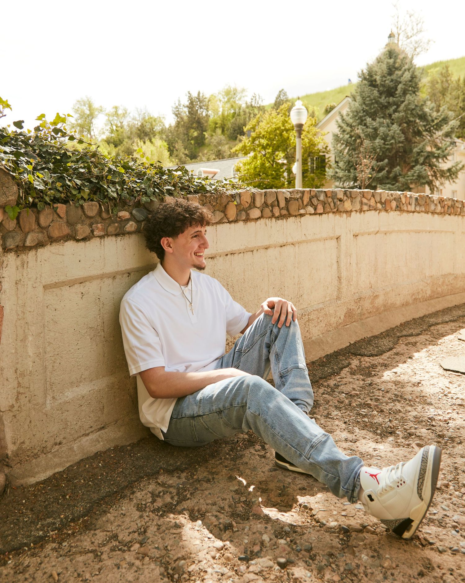 Casual portrait of someone sitting against a stone wall wearing jeans and a white shirt.