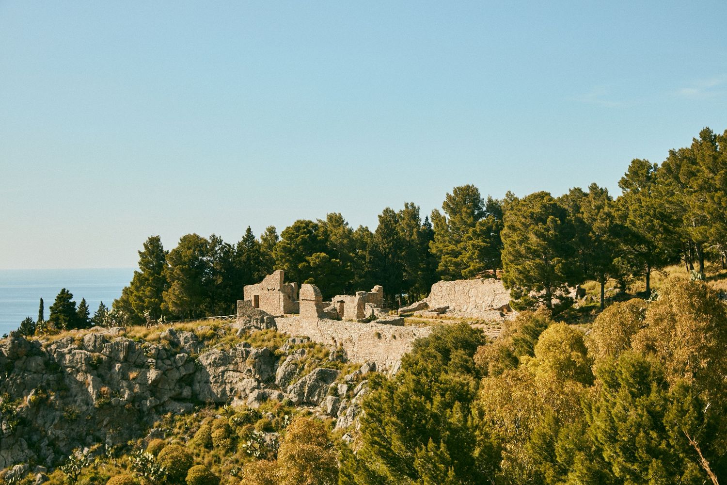 Ancient stone ruins perched on a clifftop with lush trees and Mediterranean Sea in the background.