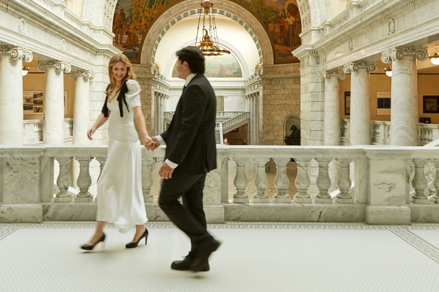 Couple in formal attire walking through ornate historic building with white columns and arches.