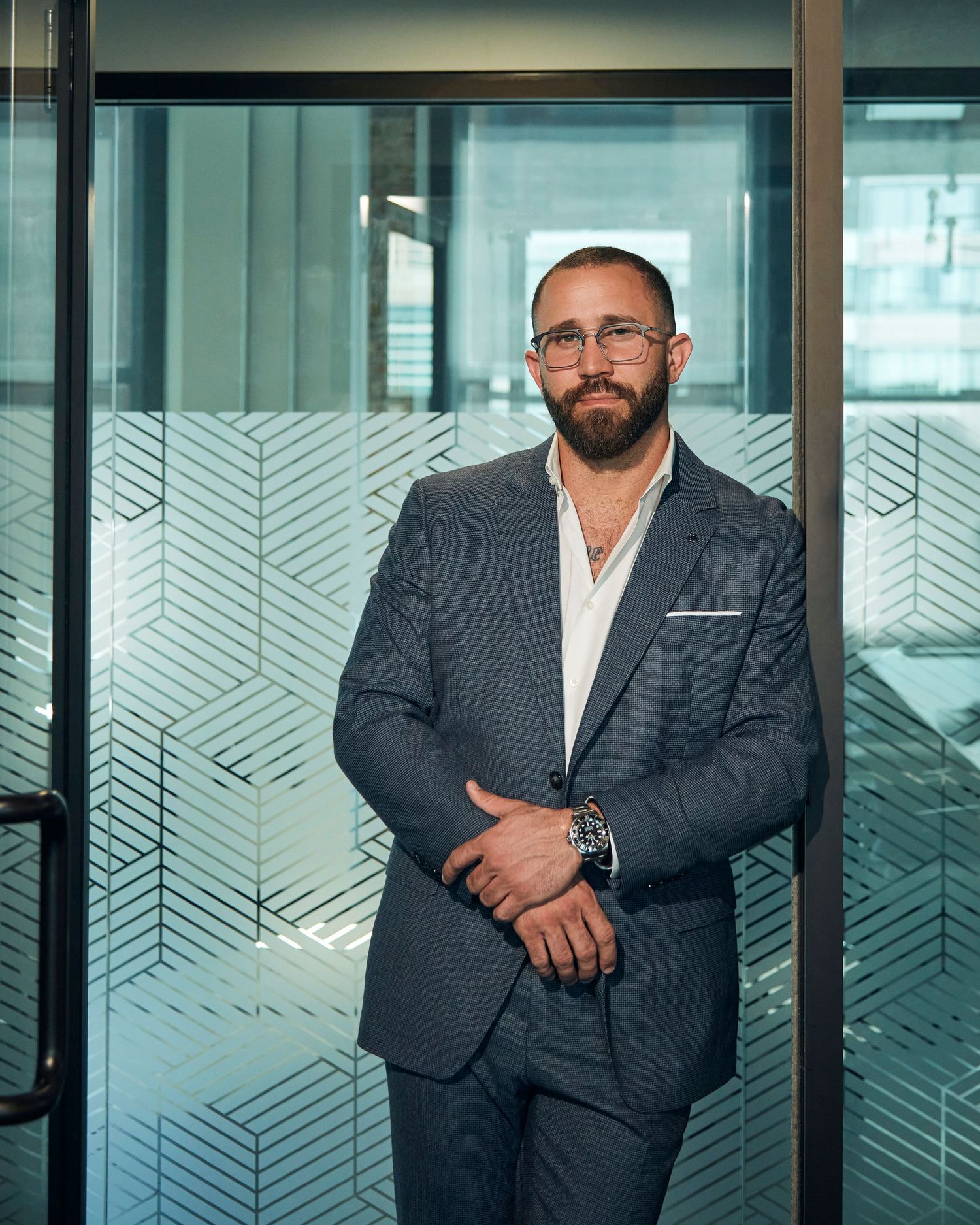 Professional in a gray suit jacket and white shirt stands confidently against frosted glass office partition.