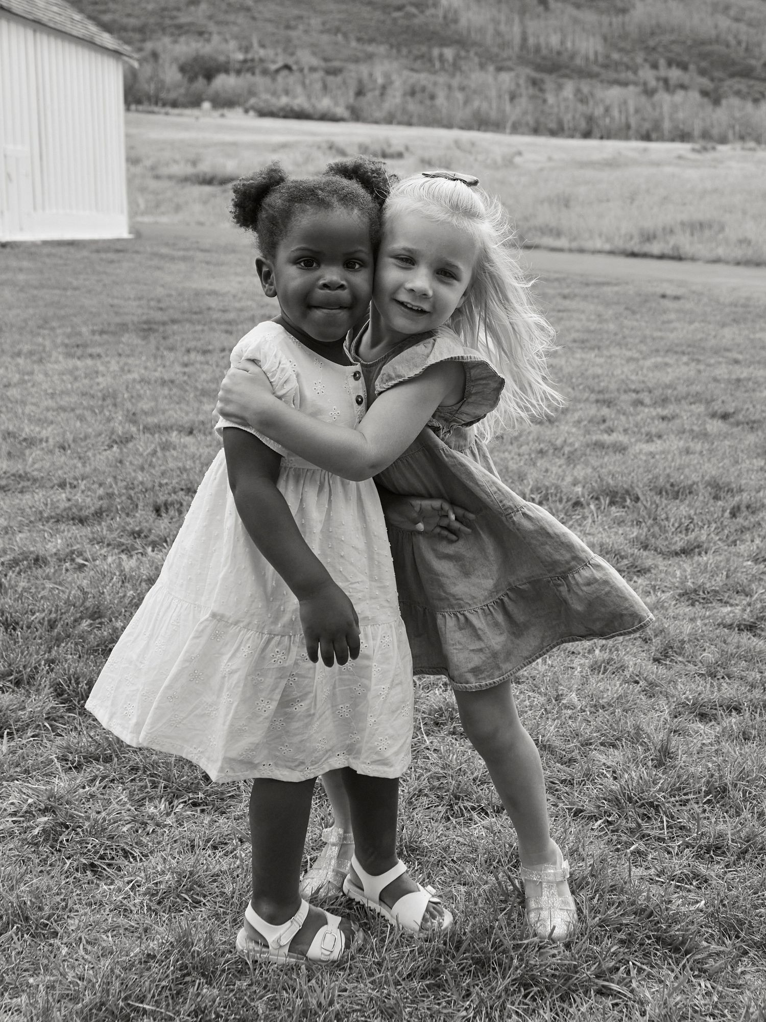 Two young friends in white dresses share a sweet embrace in black and white photography.
