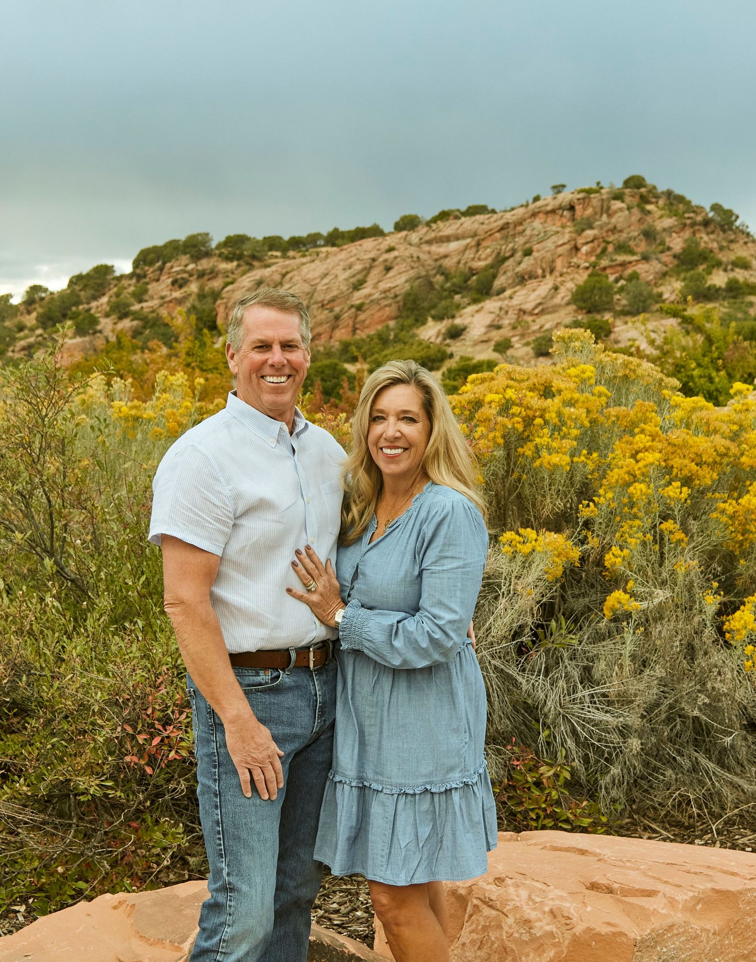 Couple poses together on red rocks with yellow wildflowers blooming in background.