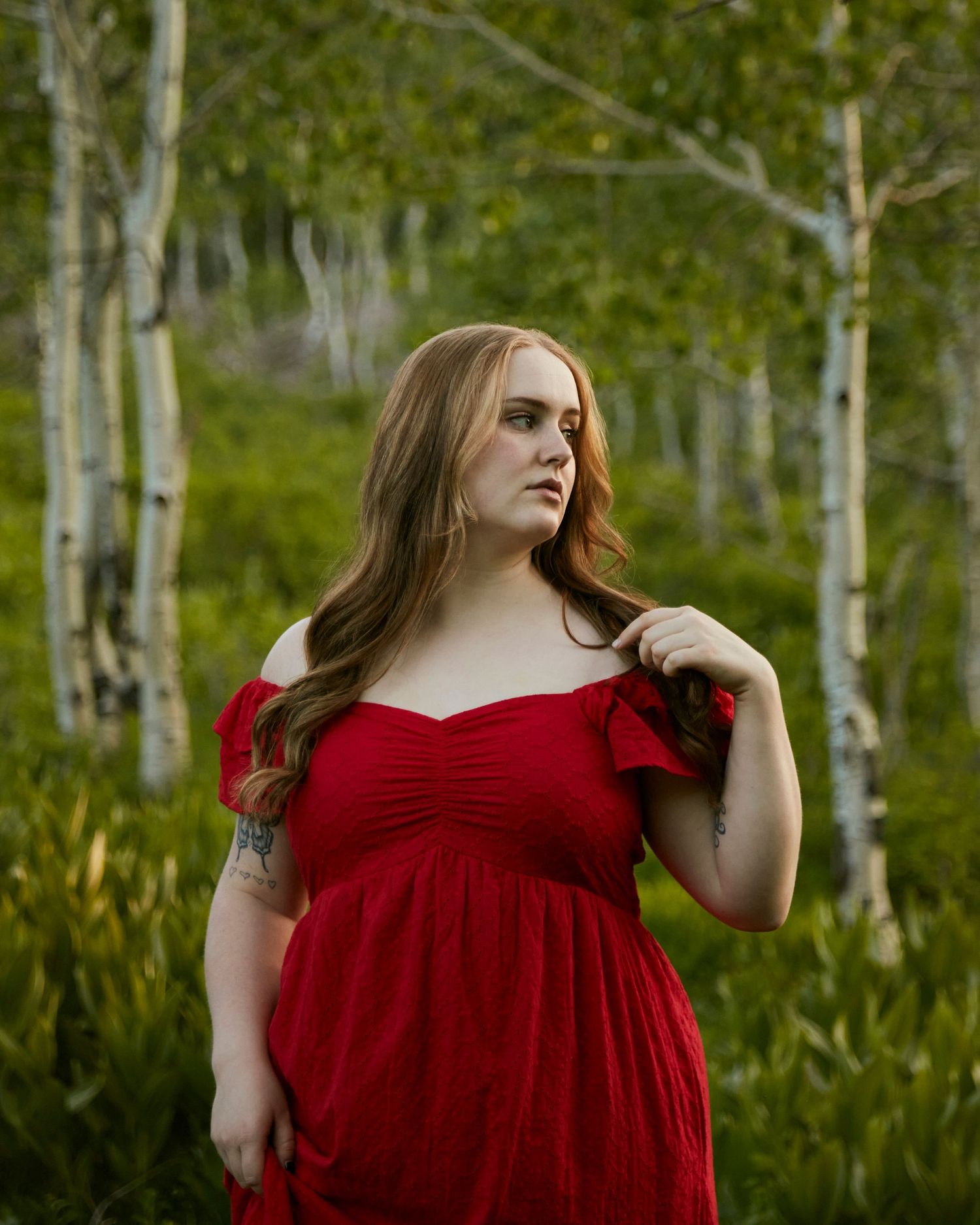 A person in a flowing red dress poses in front of a birch forest with vibrant green foliage.