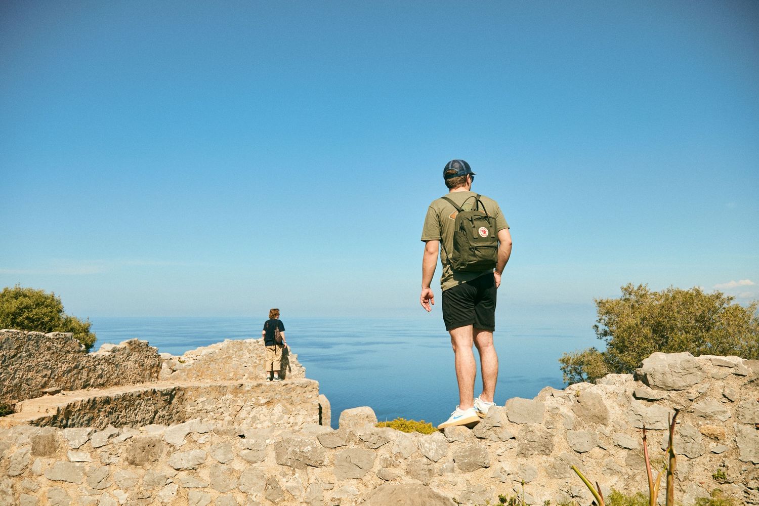 Two hikers stand on rocky cliffs overlooking a vast blue ocean on a clear sunny day.